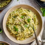 Overhead view of a rustic ceramic bowl filled with zucchini orzo, topped with finely grated Parmigiano and fresh basil leaves, with a fork resting inside. A large pan of orzo, a block of Parmigiano on a dark tray, and a second serving bowl are visible around it on a light gray surface.