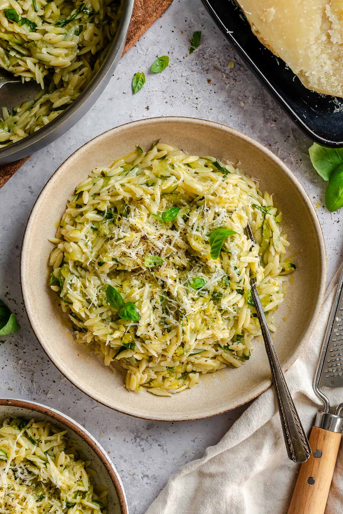 Overhead view of a rustic ceramic bowl filled with zucchini orzo, topped with finely grated Parmigiano and fresh basil leaves, with a fork resting inside. A large pan of orzo, a block of Parmigiano on a dark tray, and a second serving bowl are visible around it on a light gray surface.