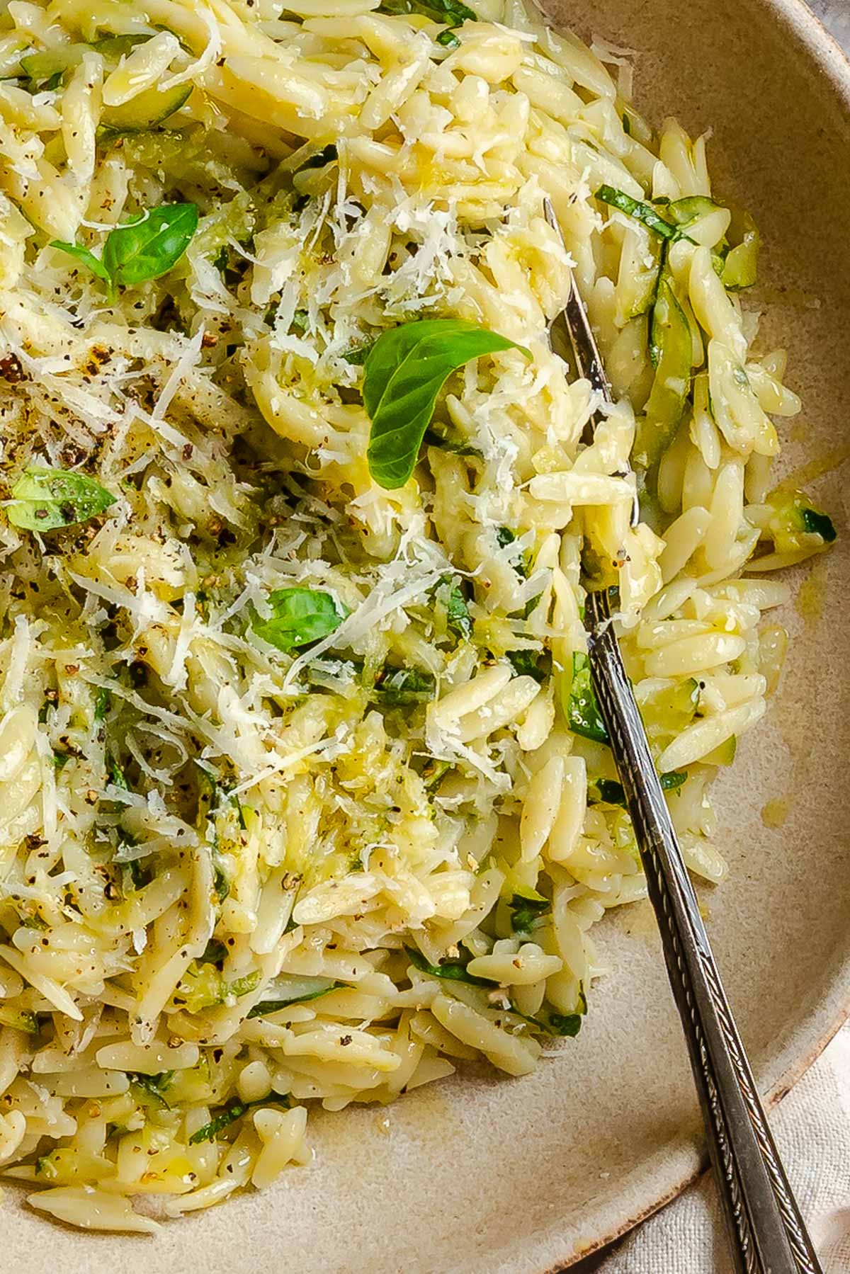 Close-up of zucchini orzo in a ceramic bowl, showing the texture of the pasta mixed with grated zucchini and shredded Parmigiano, garnished with fresh basil leaves. A fork rests at the right side of the bowl.