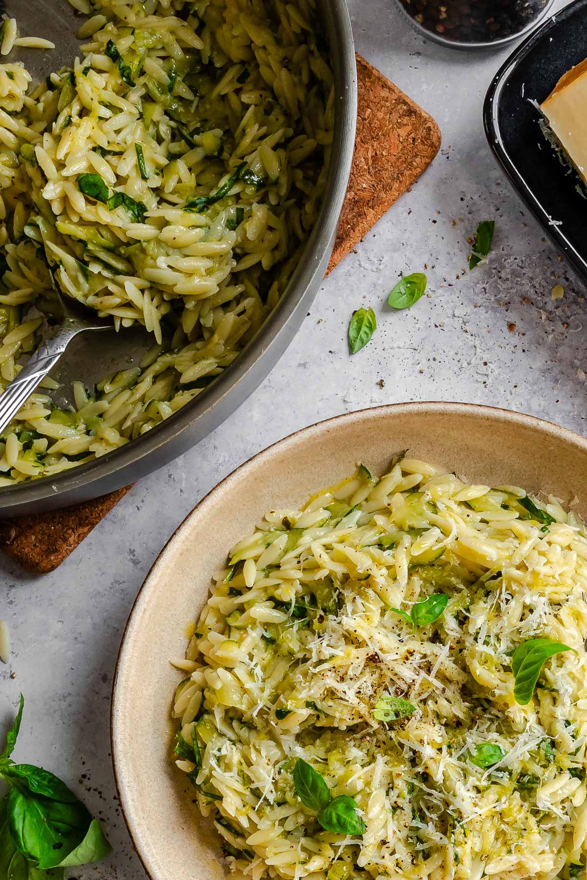 Overhead shot of a large pan of zucchini orzo on a cork trivet, with a fork resting inside, next to a ceramic serving bowl topped with Parmigiano and basil. Scattered basil leaves and a block of cheese on a dark tray are visible in the background.