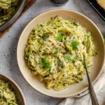 Overhead view of a ceramic bowl of zucchini orzo topped with grated Parmigiano and fresh basil, with a fork inside. Surrounding the bowl are a large pan of orzo, a second serving, fresh basil leaves, a Microplane grater, and a block of Parmigiano on a dark tray.