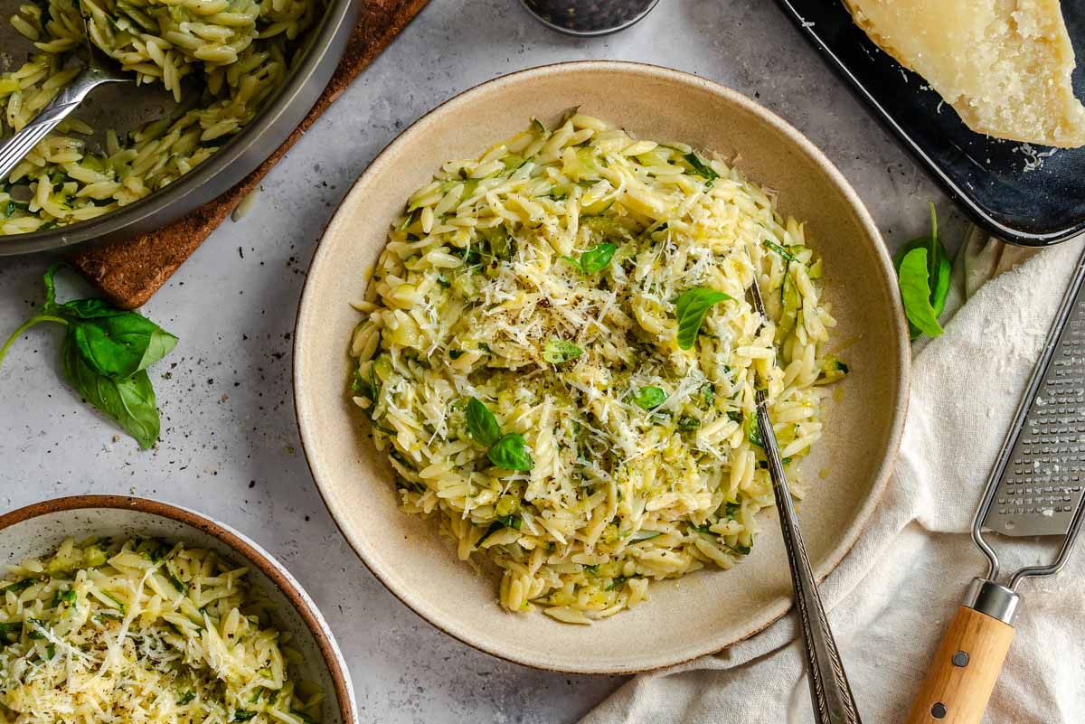 Overhead view of a ceramic bowl of zucchini orzo topped with grated Parmigiano and fresh basil, with a fork inside. Surrounding the bowl are a large pan of orzo, a second serving, fresh basil leaves, a Microplane grater, and a block of Parmigiano on a dark tray.
