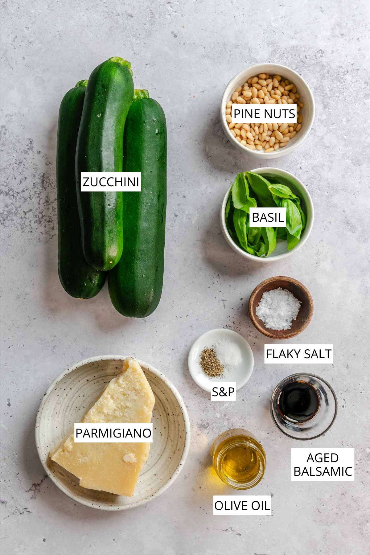 Flat lay of ingredients for grilled zucchini with balsamic and Parmigiano: two whole zucchini, a bowl of pine nuts, a bowl of fresh basil, a wedge of Parmigiano on a plate, small bowls of flaky salt and salt and pepper, a jar of olive oil, and a small glass of aged balsamic vinegar, each labeled.