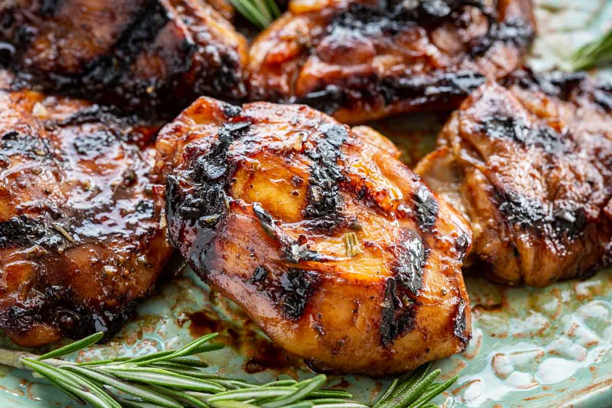 Extreme close-up of a honey balsamic chicken thigh showing the sticky, glossy glaze with caramelized bits and flecks of rosemary embedded in the char, with additional pieces and rosemary sprigs softly blurred in the background.