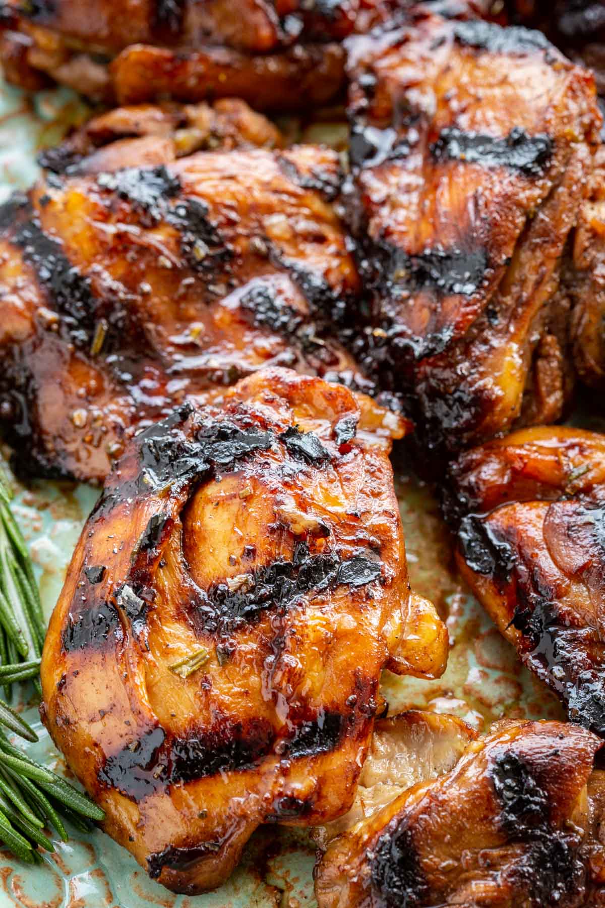 Close-up overhead shot of multiple balsamic chicken thighs on a ceramic platter, showing the deeply charred, lacquered skin glistening with reduced glaze, with a small rosemary sprig tucked between pieces.