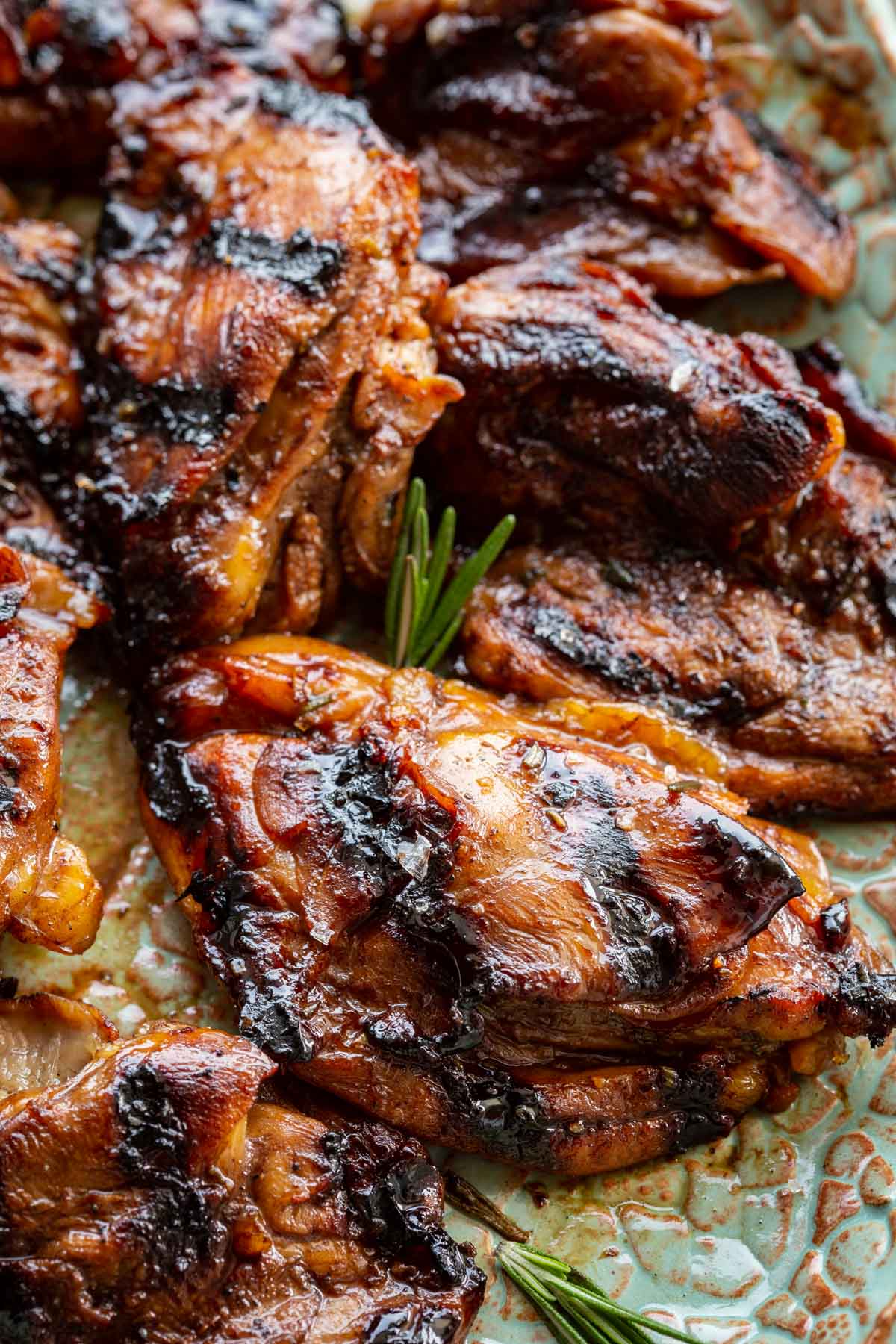 Tight close-up of balsamic grilled chicken thighs on a teal ceramic platter, highlighting the burnished, caramel-dark glaze and charred edges, with a small rosemary sprig visible between the pieces.