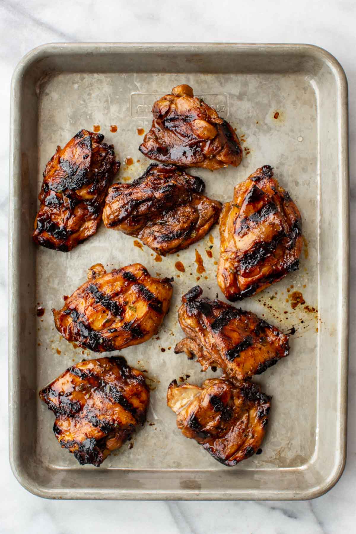 Seven balsamic-glazed grilled chicken thighs arranged on a parchment-lined aluminum sheet pan, showing deep caramelized char marks and a glossy mahogany glaze, shot overhead on a white marble surface.