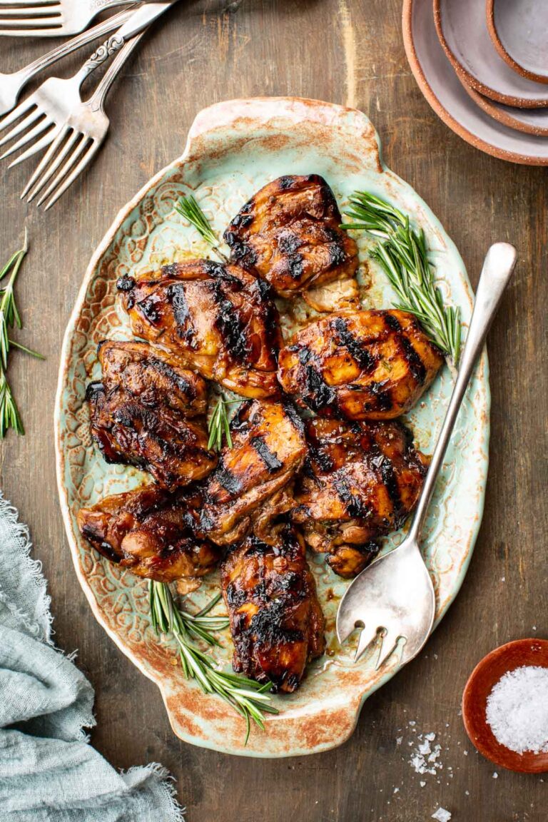 Overhead shot of balsamic grilled chicken thighs piled on a rustic oval serving platter with aged teal and terracotta glazing, garnished with fresh rosemary sprigs and a silver serving fork, surrounded by loose forks, stacked plates, and a small dish of flaky salt on a weathered wood surface.