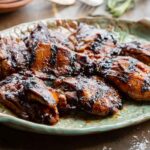 Angled close-up of honey balsamic chicken thighs on a ceramic platter, showing their deeply caramelized, lacquered surface and char marks, with fresh rosemary tucked alongside and a blurred rustic table setting in the background.