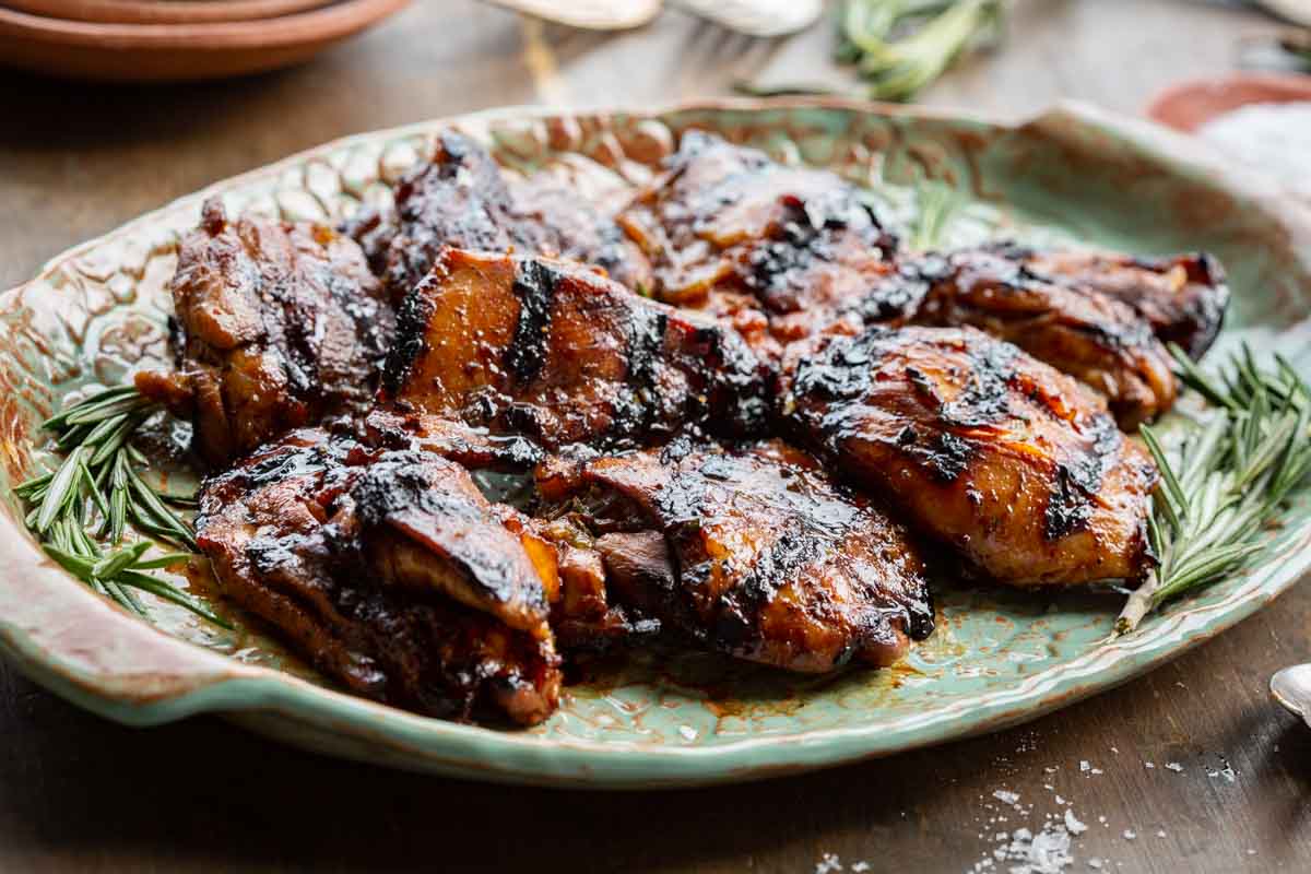 Angled close-up of honey balsamic chicken thighs on a ceramic platter, showing their deeply caramelized, lacquered surface and char marks, with fresh rosemary tucked alongside and a blurred rustic table setting in the background.
