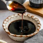 Thick balsamic reduction being poured off a spoon over a small bowl next to a kitchen towel.