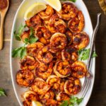 Overhead shot of blackened shrimp in a white platter next to blackening spice mix and lemon wedges.