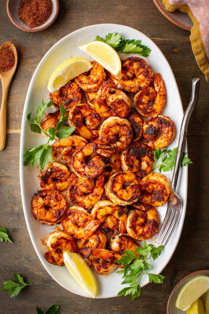 Overhead shot of blackened shrimp in a white platter next to blackening spice mix and lemon wedges.