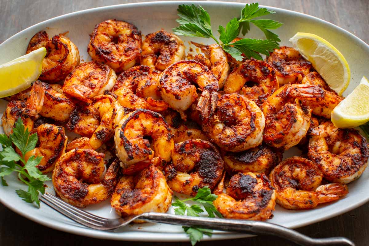 Blackened shrimp served in a large white platter with a fork and lemon wedges.