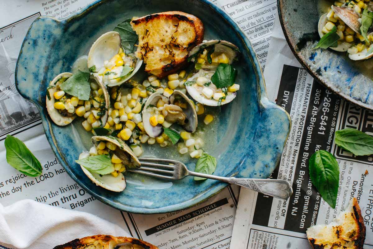 A blue ceramic bowl filled with grilled clams, corn kernels, and fresh basil leaves, served with a piece of charred grilled bread and a silver fork, set on a spread of newspaper pages.
