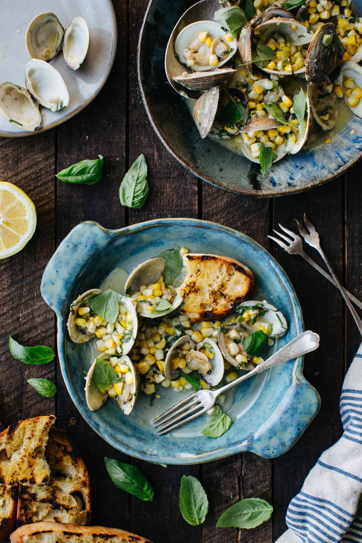 Overhead view of two blue ceramic bowls of grilled clams with corn and basil on a dark wood table, surrounded by scattered basil leaves, grilled bread slices, a halved lemon, and a striped linen napkin.