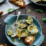 A blue ceramic bowl of grilled clams with corn and basil, shot from a slightly elevated angle, with grilled bread slices and scattered basil leaves on a dark wood surface in the background.