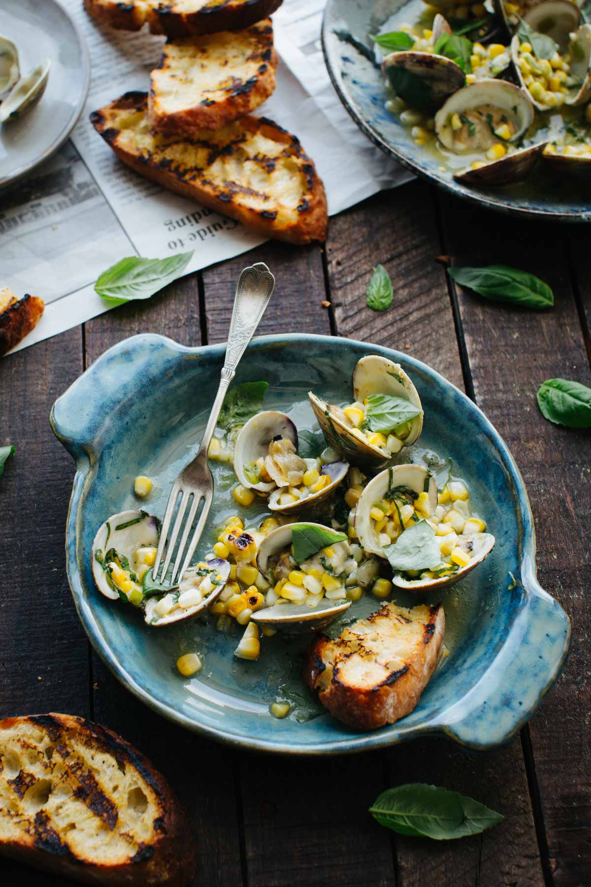 A blue ceramic bowl of grilled clams with corn and basil, shot from a slightly elevated angle, with grilled bread slices and scattered basil leaves on a dark wood surface in the background.