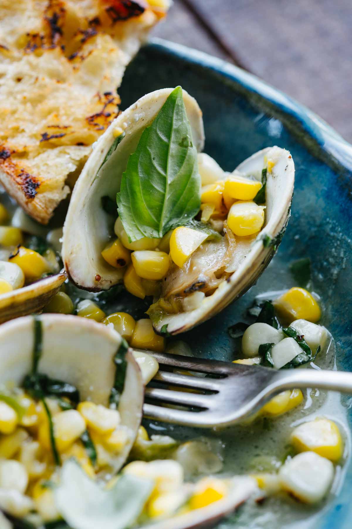 Close-up of an open clam shell filled with grilled corn kernels and topped with a fresh basil leaf, resting in a buttery broth with more clams and corn visible beneath a silver fork.