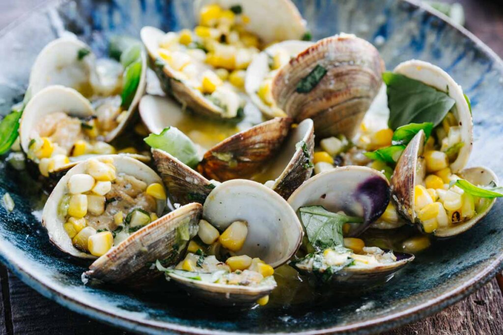 A blue ceramic bowl filled with grilled clams in their open shells, nestled in a golden butter sauce with corn kernels and fresh basil leaves scattered throughout.
