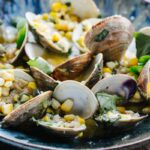 A blue ceramic bowl filled with grilled clams in their open shells, nestled in a golden butter sauce with corn kernels and fresh basil leaves scattered throughout.