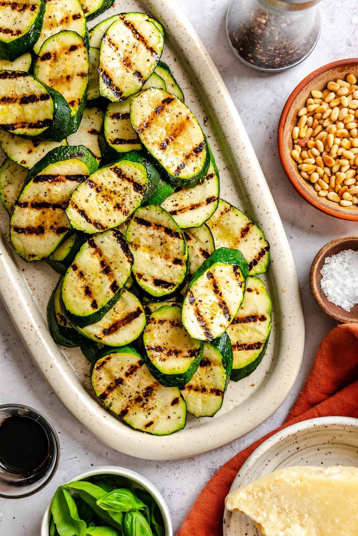A white speckled oval serving platter filled with grilled zucchini rounds topped with shaved Parmigiano, toasted pine nuts, fresh basil leaves, and a drizzle of aged balsamic vinegar, surrounded by small bowls of ingredients and an orange linen napkin.
