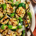 Overhead close-up of grilled zucchini rounds on a white speckled platter, topped with shaved Parmigiano, toasted pine nuts, and fresh basil, with silver serving tongs and a small wooden bowl of flaky salt on the side.