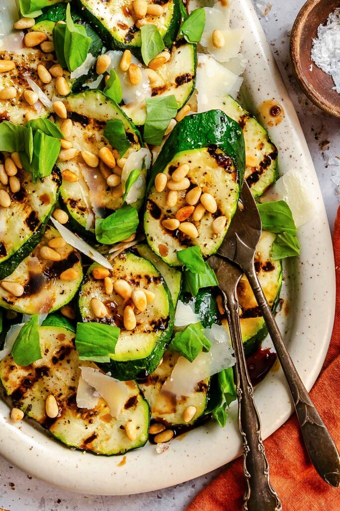 Overhead close-up of grilled zucchini rounds on a white speckled platter, topped with shaved Parmigiano, toasted pine nuts, and fresh basil, with silver serving tongs and a small wooden bowl of flaky salt on the side.