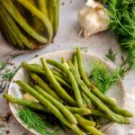 A rustic ceramic plate piled with pickled green beans, garnished with fresh dill fronds, whole garlic cloves, and scattered coriander seeds and black peppercorns. The plate sits on a light stone surface alongside a fork, a linen napkin, a head of garlic, and a partially visible jar of pickled green beans in the background. A glass jar of mixed peppercorns and fresh dill are also visible.