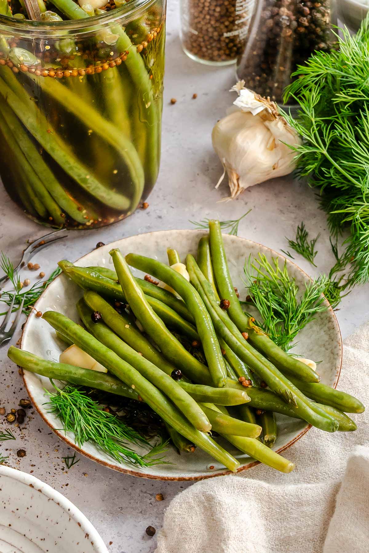 A rustic ceramic plate piled with pickled green beans, garnished with fresh dill fronds, whole garlic cloves, and scattered coriander seeds and black peppercorns. The plate sits on a light stone surface alongside a fork, a linen napkin, a head of garlic, and a partially visible jar of pickled green beans in the background. A glass jar of mixed peppercorns and fresh dill are also visible.