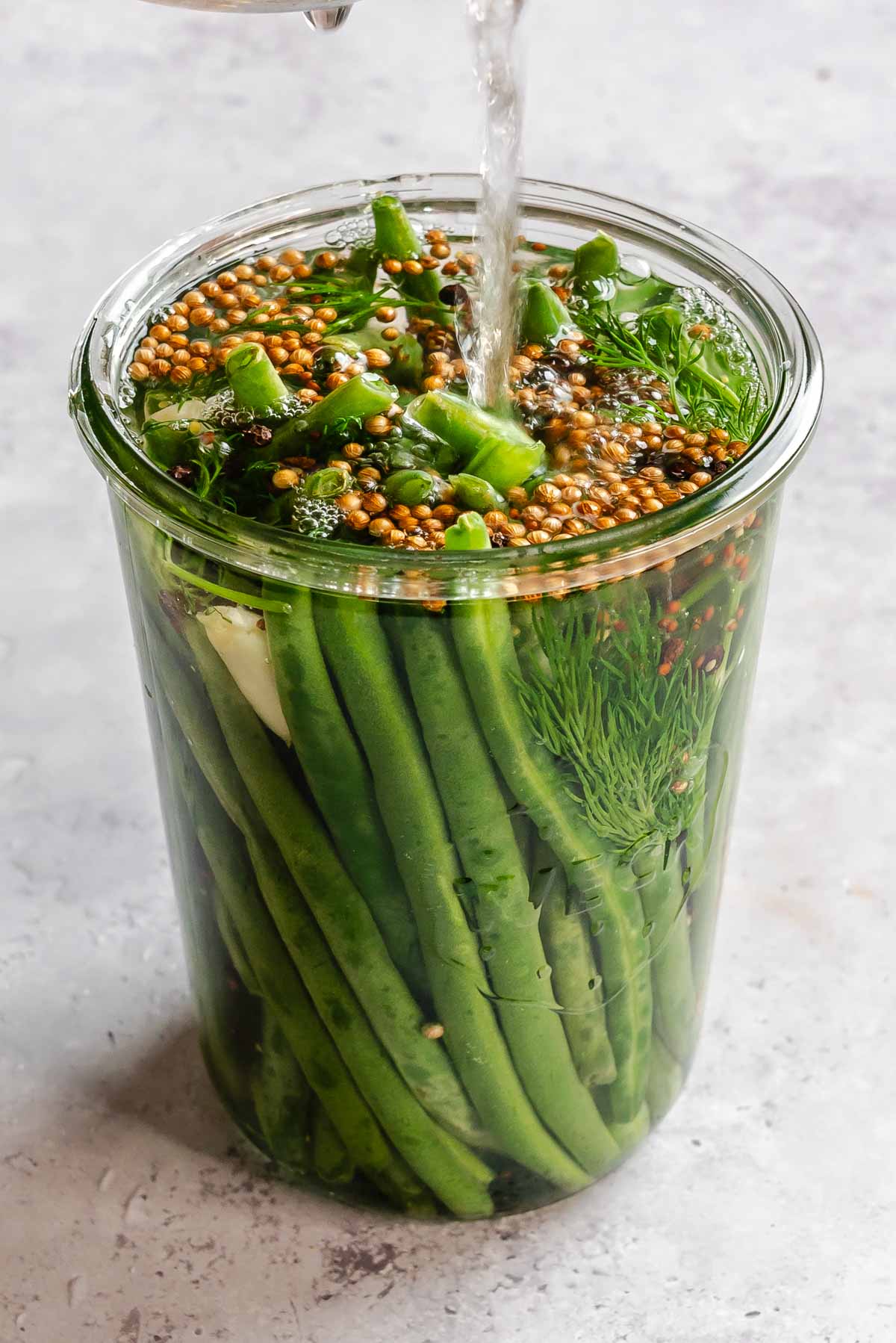 A glass jar filled with whole green beans standing upright, packed tightly and submerged in brine. Pickling liquid is being poured in from above, creating a stream of liquid that splashes over coriander seeds, fresh dill fronds, red pepper flakes, and black peppercorns floating at the surface. A slice of garlic is visible against the inside of the jar. The jar sits on a light speckled stone surface.