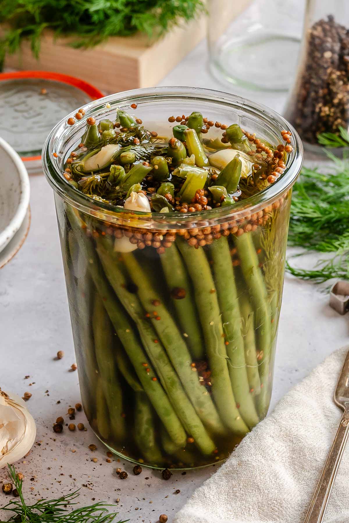 A filled Weck-style glass jar of pickled green beans, sealed and resting on a light stone surface surrounded by scattered props. The beans are packed vertically and the brine has turned slightly amber. The top of the jar is crowded with softened bean pieces, coriander seeds, garlic cloves, and wilted dill. Surrounding the jar: a linen napkin, a silver fork, scattered peppercorns, a halved garlic head, fresh dill sprigs, a jar of mixed peppercorns, and a Weck lid with orange rubber seal.