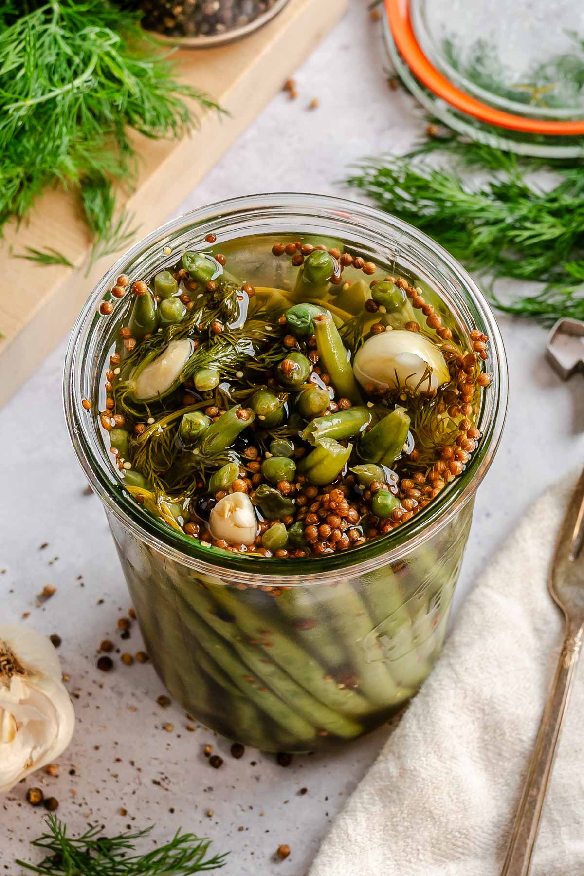 An overhead close-up of a glass jar filled with pickled green beans, viewed from directly above. The top of the jar shows a jumble of softened bean tips, whole garlic cloves, coriander seeds, wilted dill, and black peppercorns submerged in amber brine. A wooden cutting board, fresh dill, and a jar of peppercorns are visible in the background.