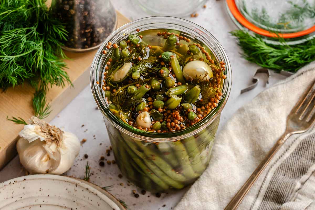 A slightly elevated overhead shot of a glass jar packed with pickled green beans, garlic cloves, dill, coriander seeds, and peppercorns in amber brine. A whole garlic bulb sits to the left of the jar on a wooden board, and fresh dill sprigs, a silver fork, a linen napkin, and a jar of mixed peppercorns are arranged around it on a light stone surface.