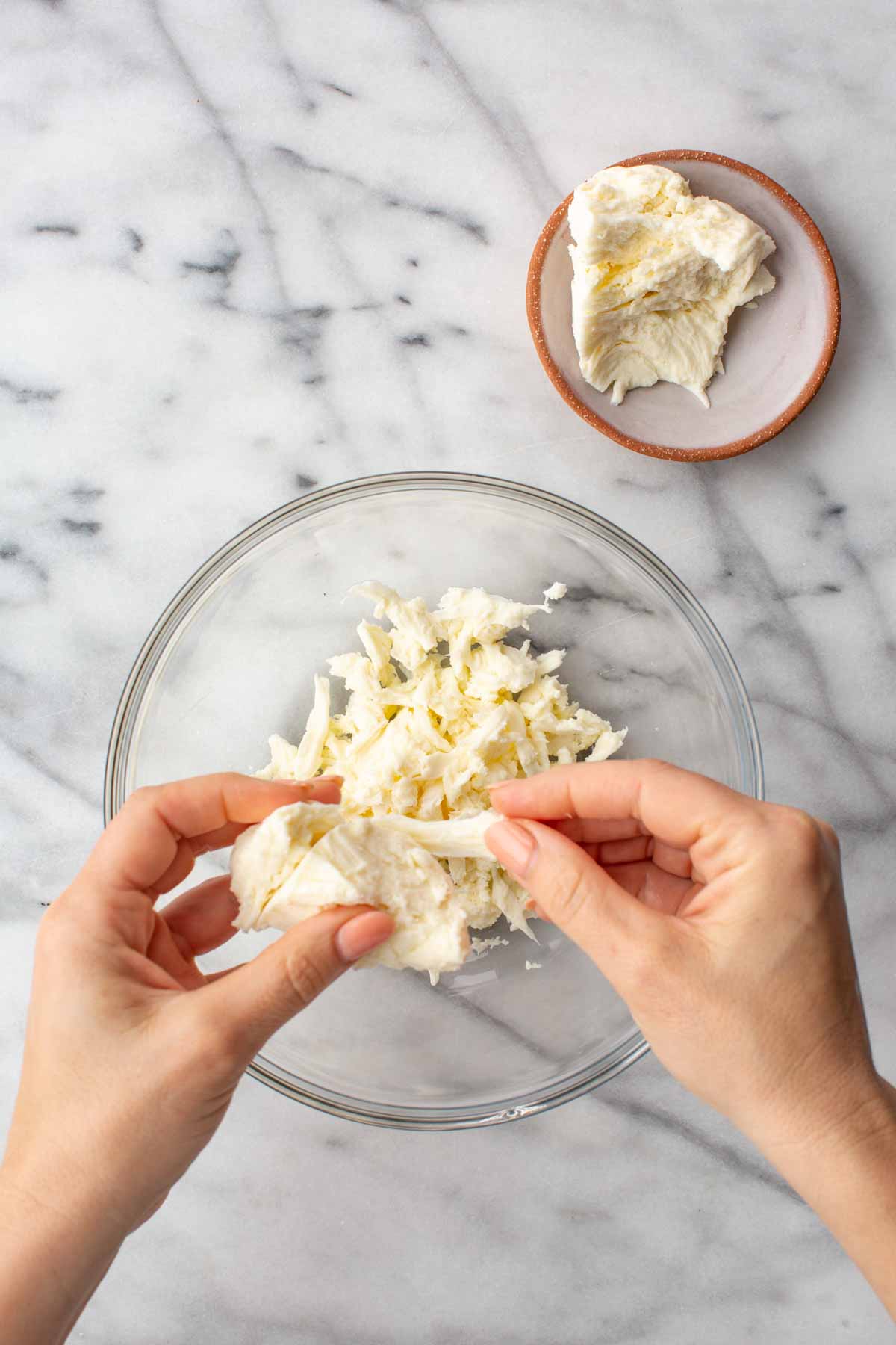 Hands shredding a piece of fresh mozzarella into thin strands over a glass bowl already filled with torn mozzarella curds.