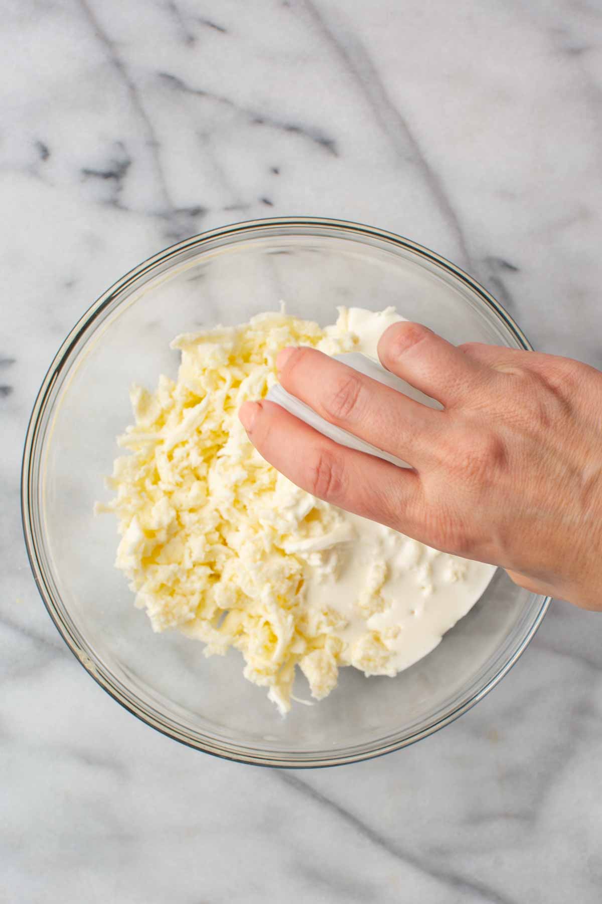A hand pressing and mixing shredded mozzarella into heavy cream in a glass bowl, combining the curds with the liquid.
