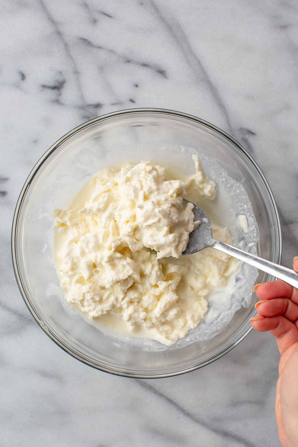 A spoon lifting a portion of creamy mozzarella curds from a glass bowl, showing the rich, soft texture of the mixed stracciatella.