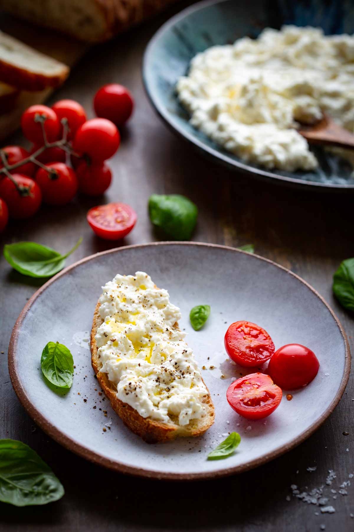 A slice of toasted bread topped with stracciatella cheese, olive oil, and black pepper on a plate, with fresh basil leaves and halved cherry tomatoes alongside, and a bowl of stracciatella in the background.