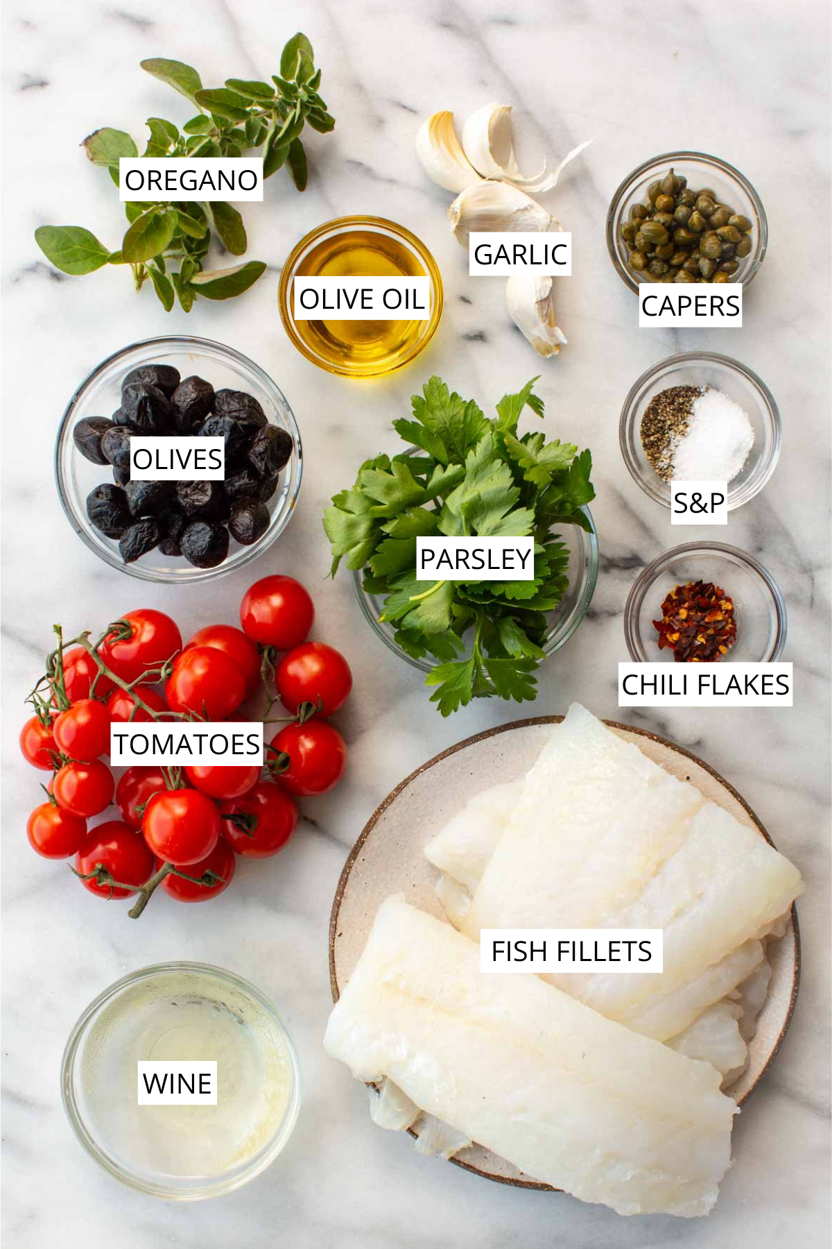 Labeled ingredients for fish acqua pazza laid out on a marble surface, including oregano, garlic, olive oil, capers, black olives, parsley, salt and pepper, chili flakes, cherry tomatoes, white fish fillets, and white wine.