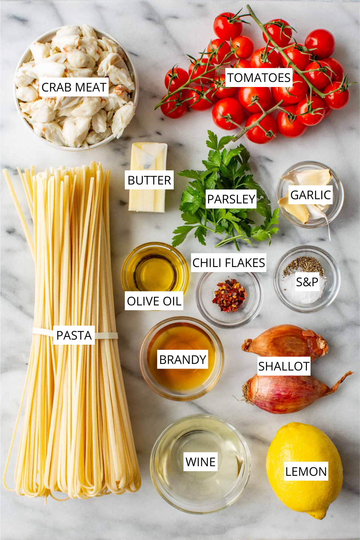 Labeled ingredients for crab pasta: crab meat, tomatoes, pasta, butter, parsley, garlic, olive oil, chili flakes, salt and pepper, brandy, shallot, wine, and lemon.