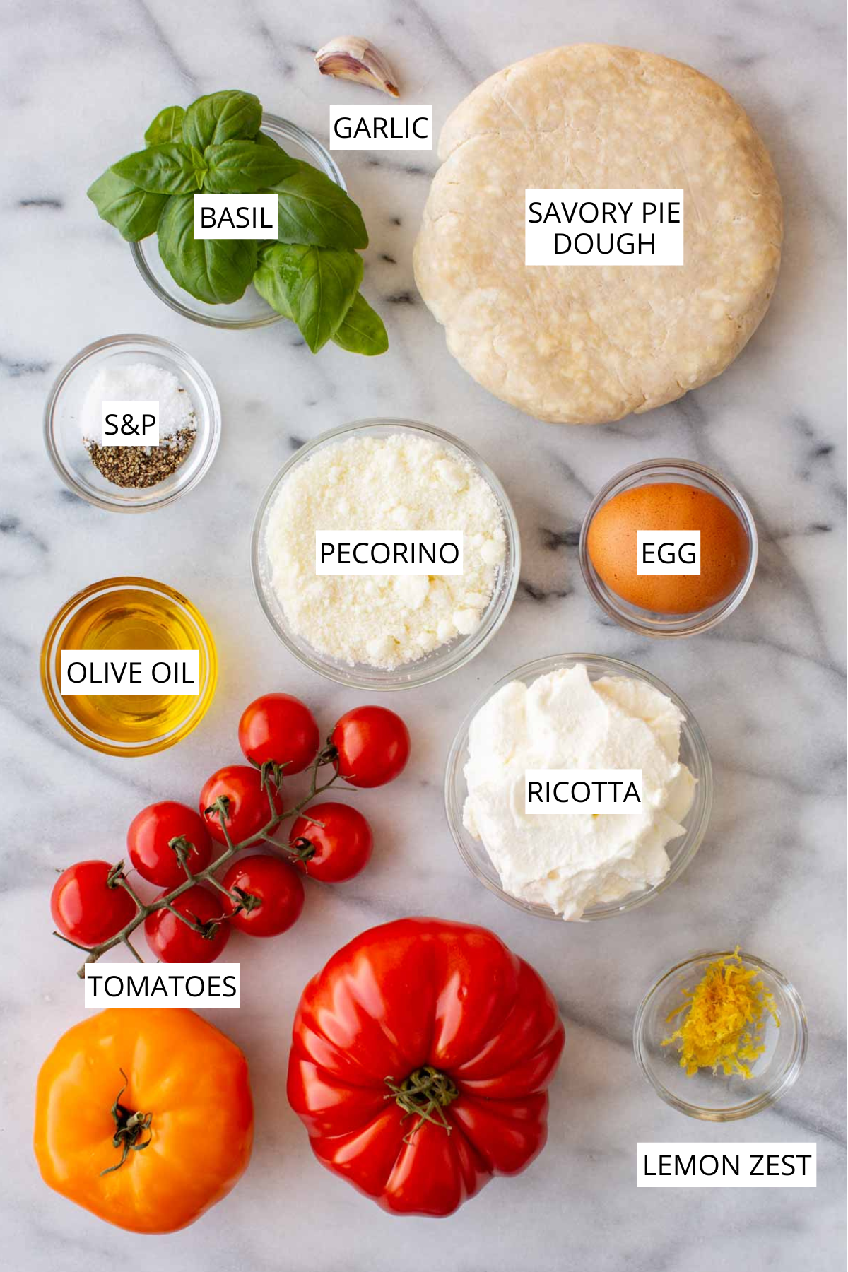 Overhead flat-lay of labeled crostata ingredients, including savory pie dough, basil, garlic, salt and pepper, pecorino, egg, olive oil, ricotta, heirloom and cherry tomatoes, and lemon zest, arranged on a marble surface.