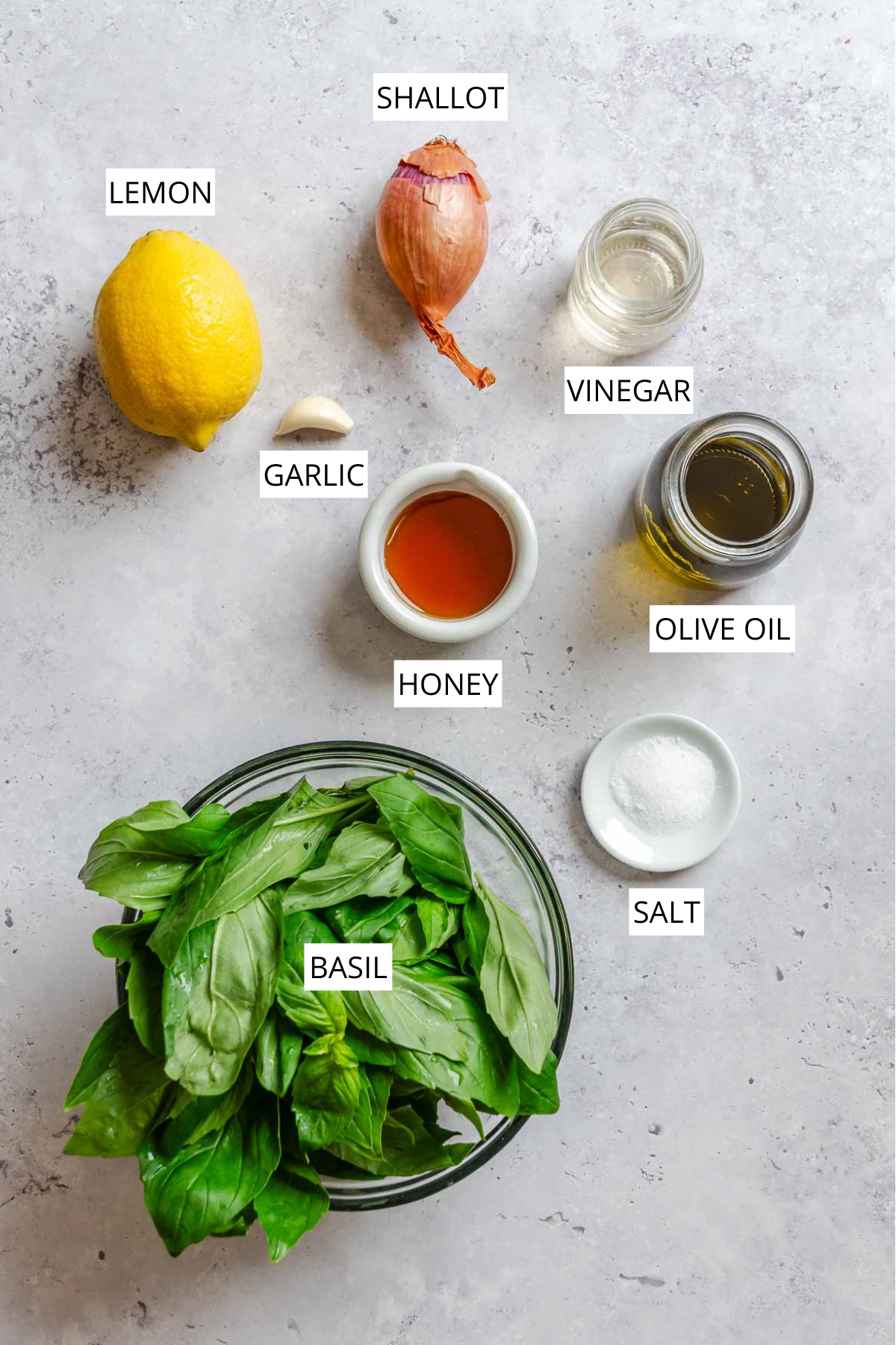 Overhead shot of basil vinaigrette ingredients labeled on a gray surface, including a lemon, shallot, vinegar, garlic, olive oil, honey, salt, and a bowl of fresh basil leaves.