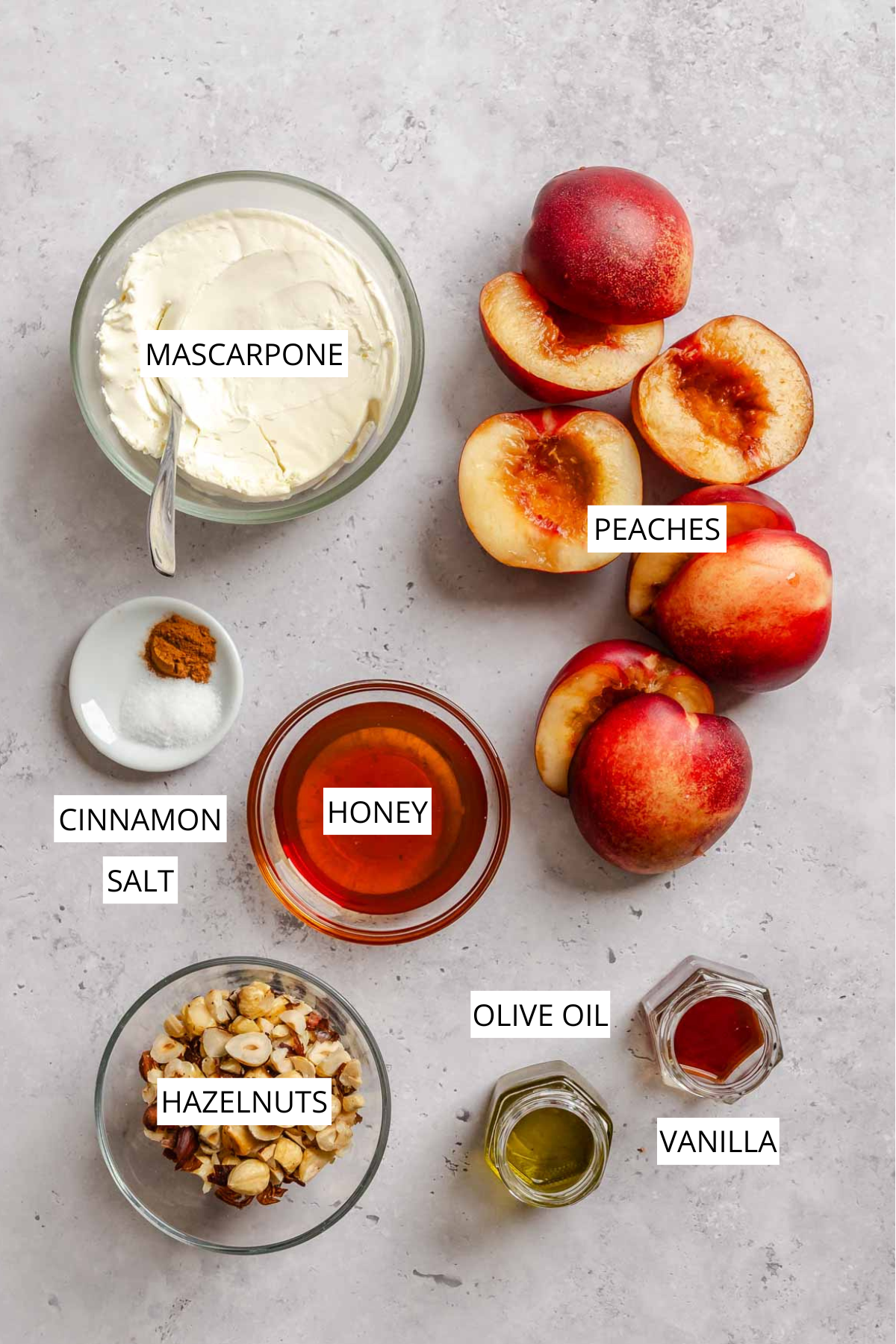 Flat-lay of labeled ingredients for grilled peaches with mascarpone: mascarpone, peaches, cinnamon, salt, honey, hazelnuts, olive oil, and vanilla, each in small glass bowls on a gray surface.