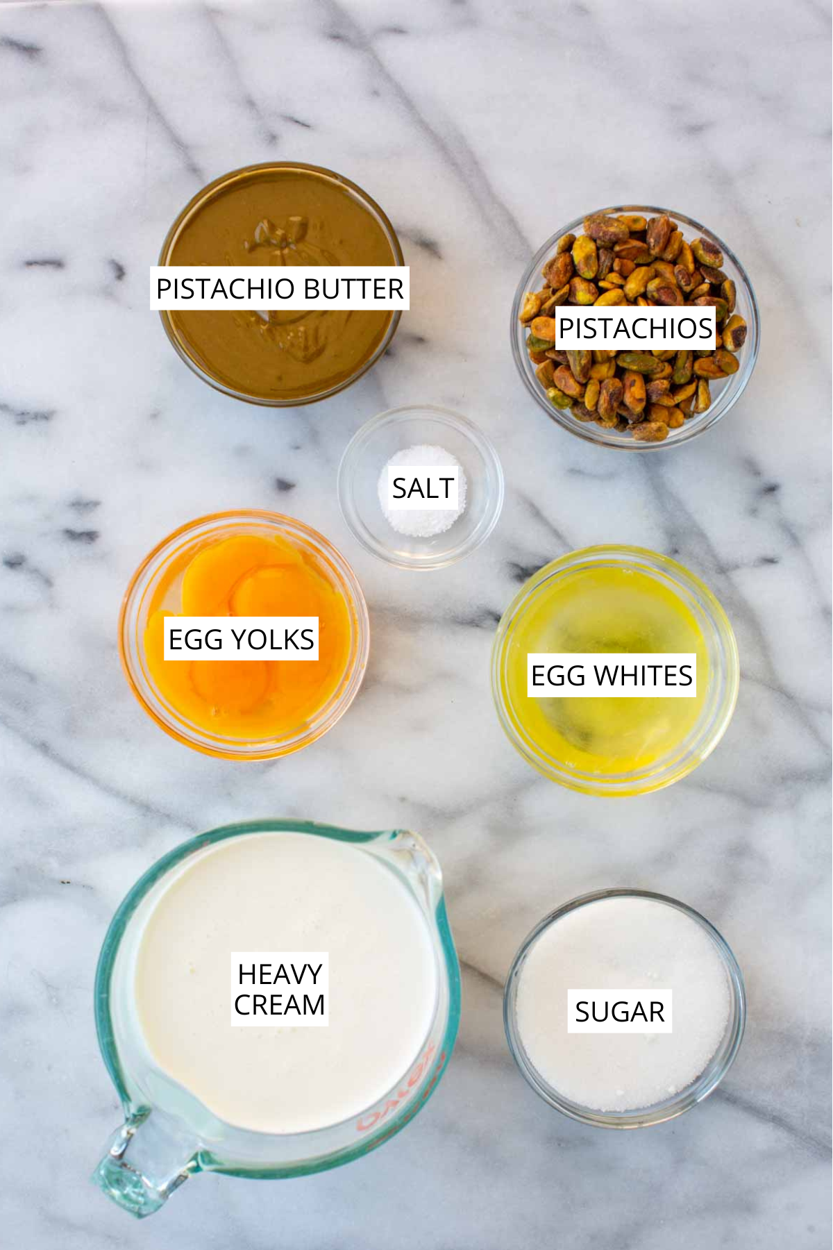 Overhead shot of labeled pistachio semifreddo ingredients on a marble surface: pistachio butter, whole pistachios, salt, egg yolks, egg whites, heavy cream, and sugar.