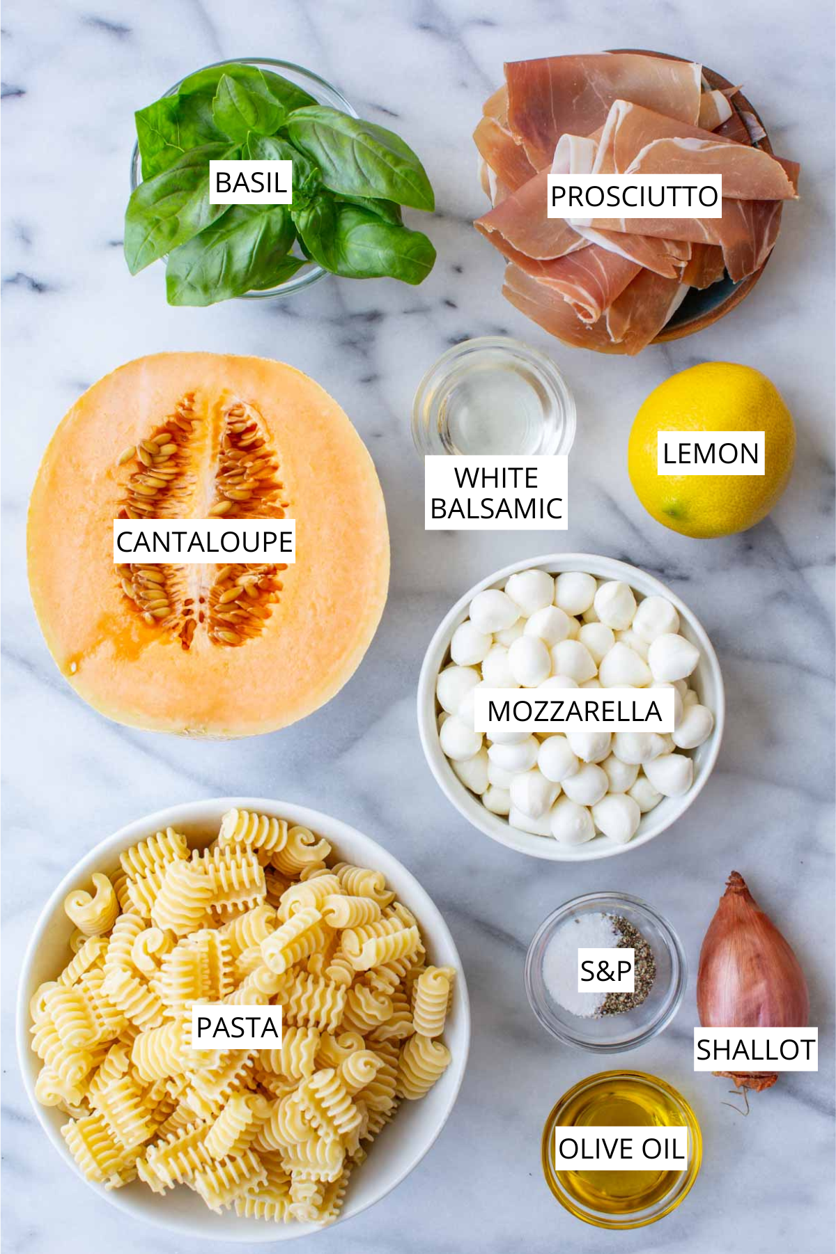 Overhead flat lay of labeled ingredients for prosciutto melon pasta salad: basil, prosciutto, cantaloupe, white balsamic vinegar, lemon, mozzarella, pasta, salt and pepper, shallot, and olive oil, arranged on a marble surface.