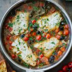 Overhead shot of fish acqua pazza in a stainless steel skillet, with four white fish fillets simmered in a tomato and olive broth, garnished with fresh parsley, surrounded by cherry tomatoes, oregano, and toasted bread.