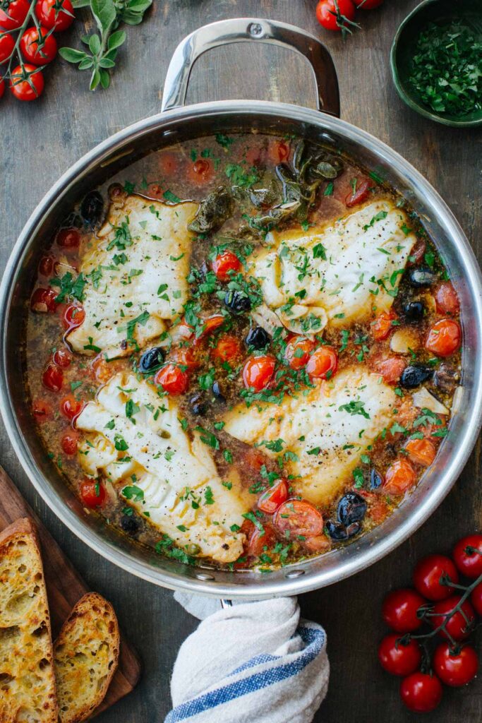 Overhead shot of fish acqua pazza in a stainless steel skillet, with four white fish fillets simmered in a tomato and olive broth, garnished with fresh parsley, surrounded by cherry tomatoes, oregano, and toasted bread.