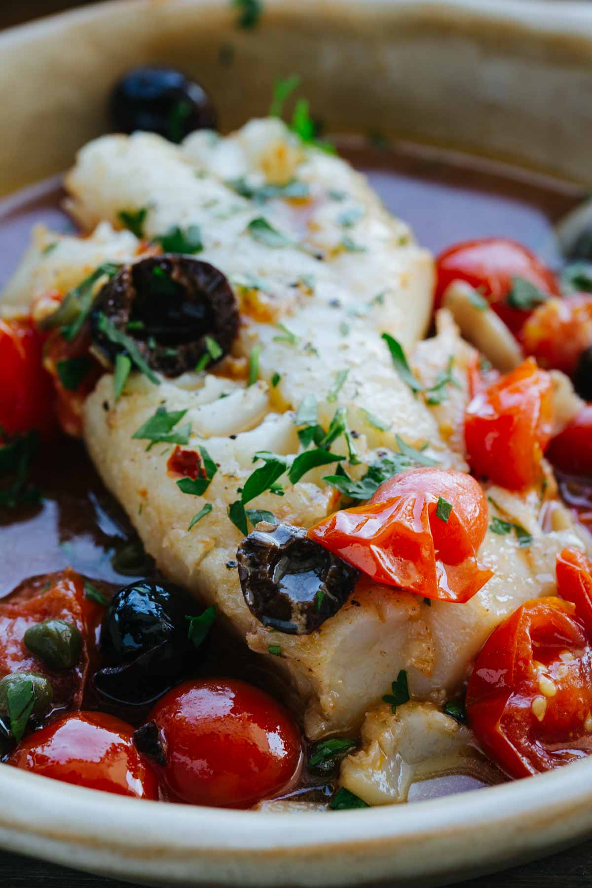 Close-up of a single fish fillet in acqua pazza broth, topped with chopped parsley, black olives, and burst cherry tomatoes in a ceramic bowl.