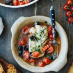 Bowl of fish acqua pazza with a fork resting inside, surrounded by cherry tomatoes, olives, and toasted bread, with the full skillet visible in the background.