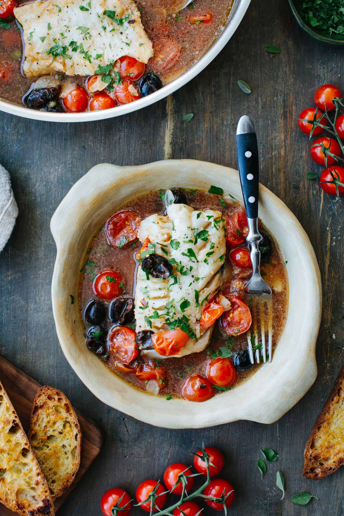 Bowl of fish acqua pazza with a fork resting inside, surrounded by cherry tomatoes, olives, and toasted bread, with the full skillet visible in the background.